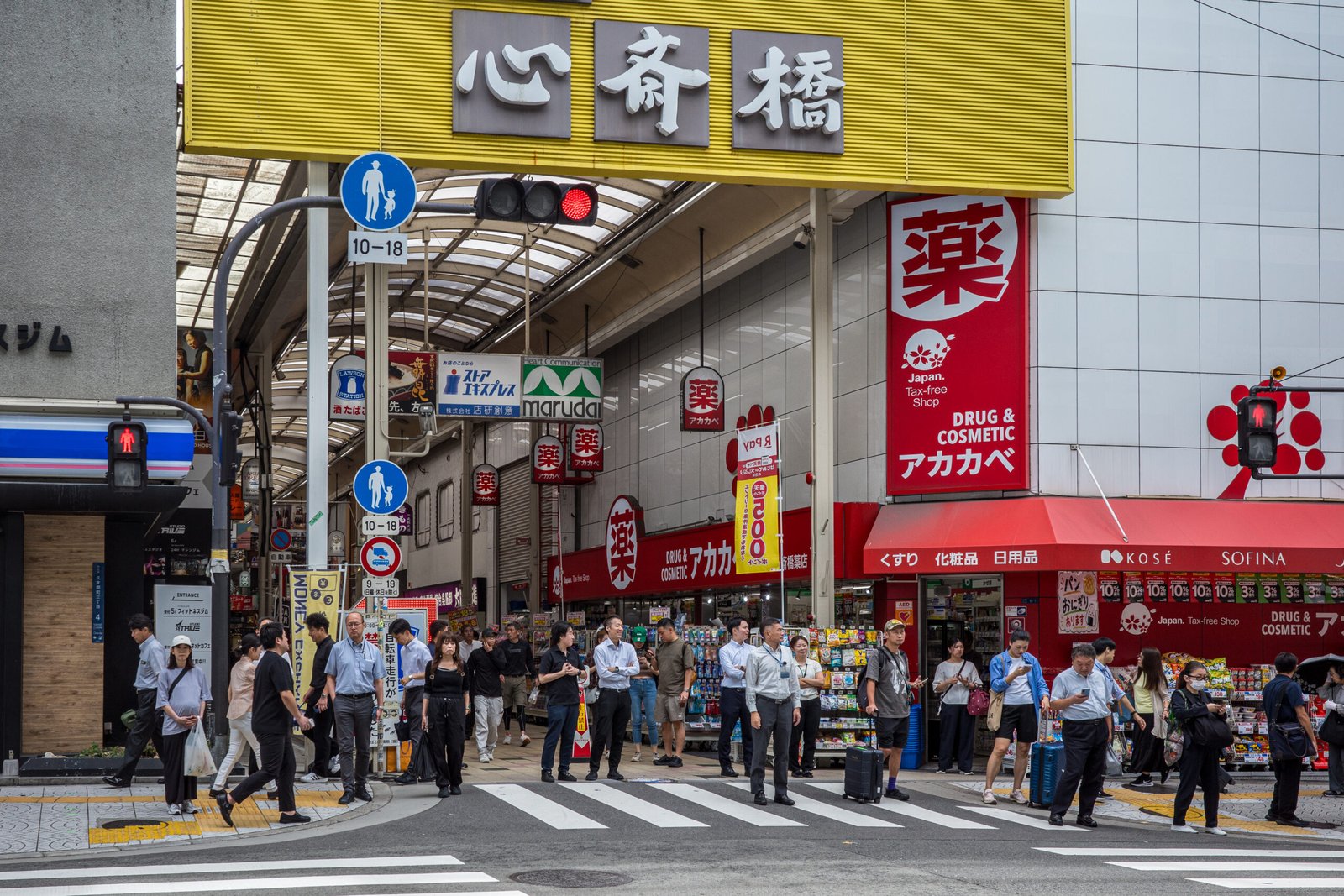 A busy intersection in Osaka with many pedestrians waiting to cross, several pushing luggage.