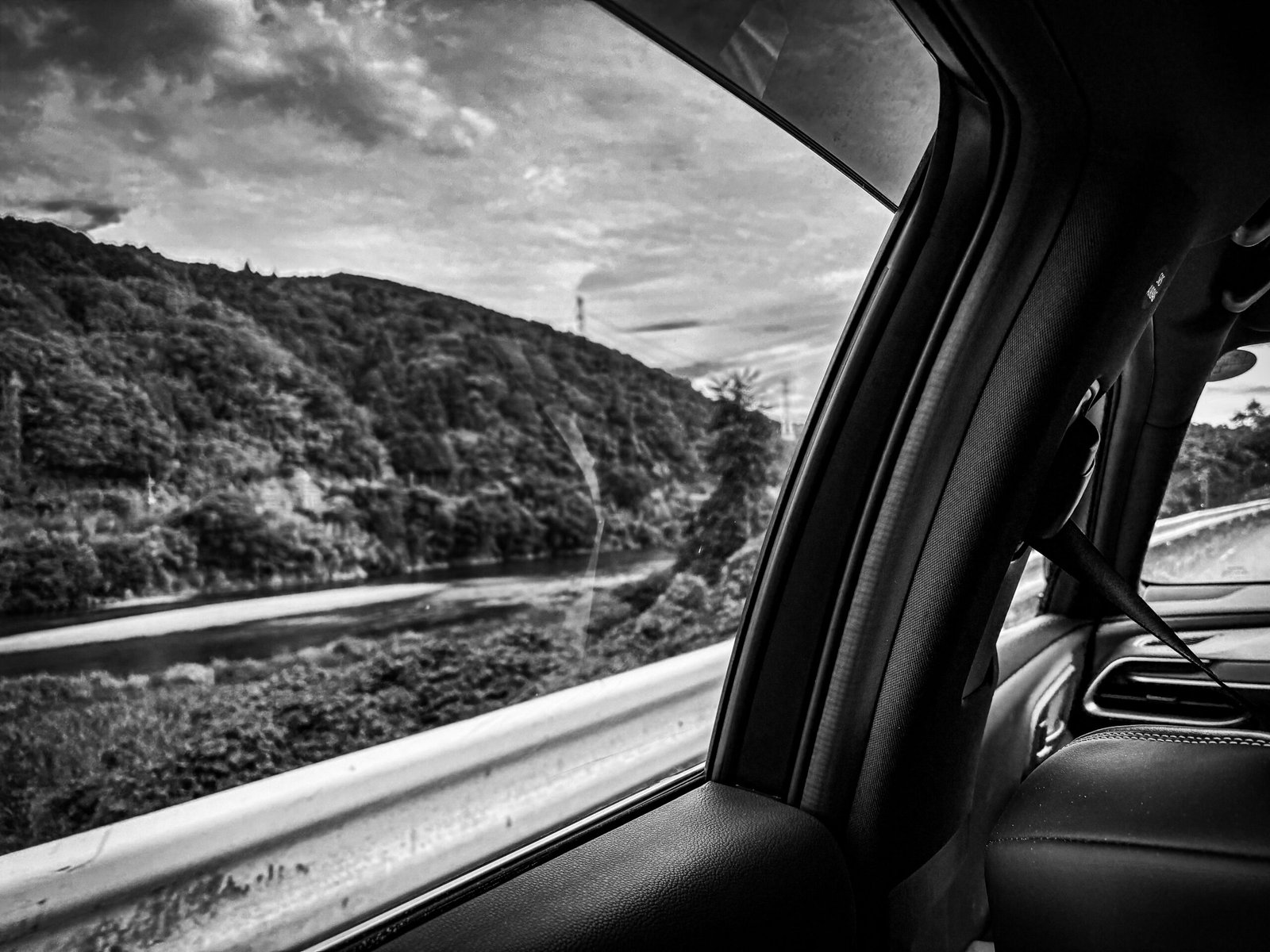 A black and white image of the view from a car window, showing a landscape with a river, mountains and clouds.
