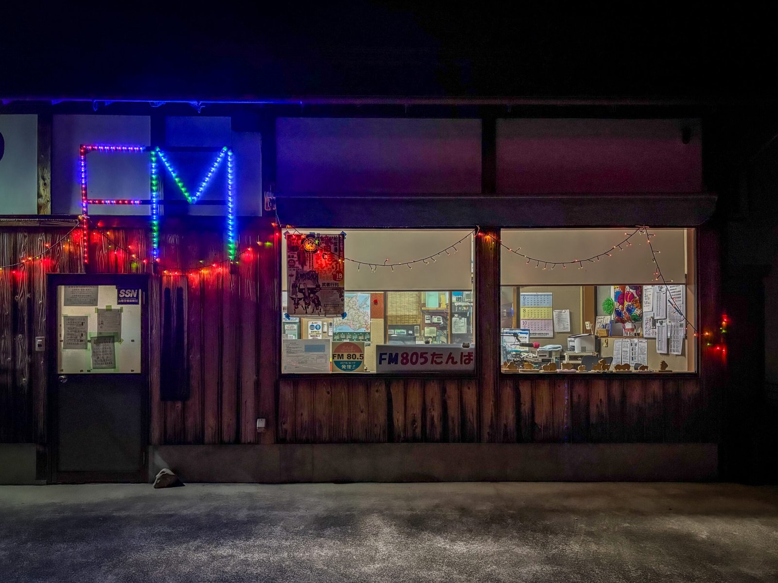 A night view outside of a radio studio, with neon lights forming the letters "FM," and a sign "FM 80.5 Tamba" in the window.