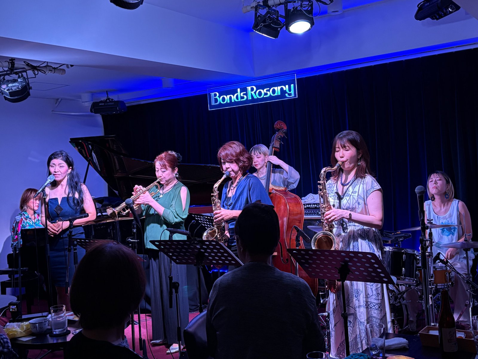 Seven female musicians playing jazz instruments on a stage, with audience members in the foreground, and a sign saying "Bonds Rosary" in the background.